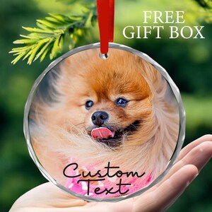May include: Round glass ornament with a photo of a small, fluffy, light brown dog with a pink tongue sticking out. The ornament has a silver rim and a red ribbon attached to the top. The text "Custom Text" is printed on the ornament.