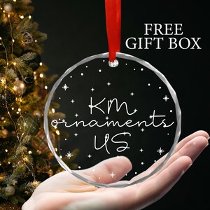 May include: A clear glass Christmas ornament with the text "KM ornaments US" and white snowflakes on a white background.