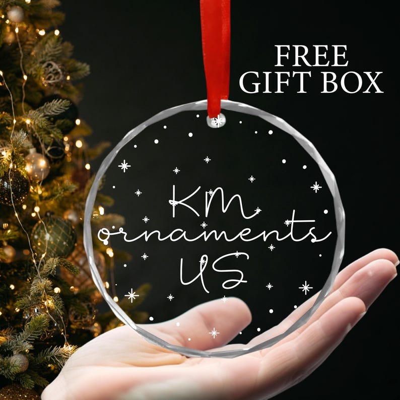 May include: A clear glass Christmas ornament with the text "KM ornaments US" and white snowflakes on a white background.