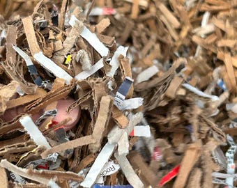 May include: Close-up of shredded cardboard and paper, creating a textured, brown and white mix. Some pieces have printed text and colorful accents. The image shows a close-up of the shredded material.