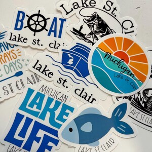 May include: A collection of stickers with lake-themed designs. The stickers feature text like "Lake St. Clair" and "Lake Life", along with images of a boat wheel, fish, and a sunrise over water. Colors include blue, orange, and white.