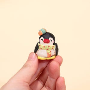 Könnte beinhalten: Handgefertigte Pinguin-Figur. Der Pinguin hat einen schwarzen Kopf und Flügel, einen weißen Bauch und einen roten Schnabel. Er trägt einen grünen Schal mit gelben Punkten und einen kleinen Hut. Die Figur ist ca. 4 cm groß.