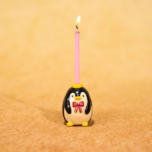 Puede incluir: Un original portavelas con forma de pingüino con una vela rosa encendida. El pingüino tiene un cuerpo negro, vientre blanco y una pajarita roja. La vela está insertada en un anillo amarillo en la cabeza del pingüino. El fondo es de color beige.