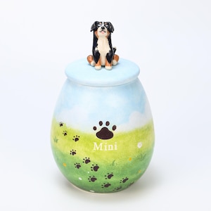 Pode incluir: Uma urna de cerâmica com uma paisagem pintada de um campo verde e céu azul. Uma figura de cão preto, castanho e branco está no topo da tampa azul clara. A palavra "Mini" está impressa na urna.