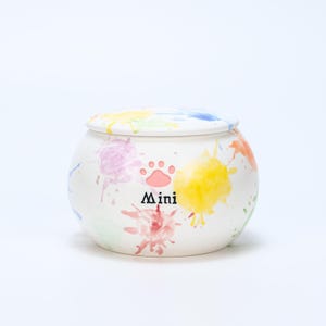May include: A white ceramic pet urn with a lid, decorated with colorful paint splatters in shades of pink, yellow, blue, and orange. The urn features a paw print design and the word "Mini" in black lettering.