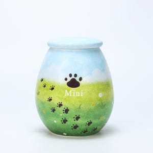 Pode incluir: Uma pequena urna de cerâmica em forma de ovo com tampa azul clara. A urna é pintada com um design de paisagem verde e azul, com pegadas e a palavra "Mini". A urna é projetada para cremação de animais de estimação.