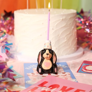 May include: A ceramic dog-shaped candle holder with a lit pink candle. The black and white dog has a pink heart on its chest and a party hat. A white cake with pearl-like decorations is in the background.