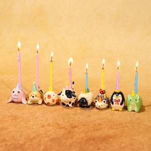 May include: A collection of eight lit birthday candles. Each candle is set atop a different animal figurine, including a seal, dog, cow, sheep, cat, penguin, and frog. The candles are various colors, and the figurines are detailed.