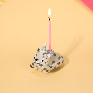 以下が含まれることがあります： 点灯したピンク色のろうそくが付いた、セラミック製のユキヒョウの誕生日キャンドルホルダー。ユキヒョウは灰色の斑点、青いパーティーハット、笑顔の顔をしています。ろうそくはユキヒョウの背中に配置されています。