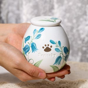 May include: White ceramic pet urn with a lid, decorated with blue floral patterns and a brown paw print. The urn has the word "Scritchy" written on it. The urn is designed to hold the ashes of a beloved pet.