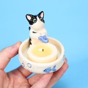 May include: A ceramic candle holder featuring a black and white cat figurine holding a blue fish. The holder has a cream-colored bowl with blue fish and bubble designs. A lit yellow candle sits inside.