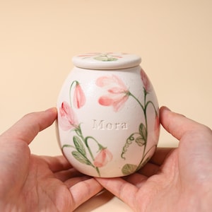 May include: A cream-colored ceramic jar with a matching lid, featuring hand-painted pink sweet pea flowers and green vines. The word "Mora" is embossed in the center of the jar. The vessel has a rounded, egg-like shape with a smooth, matte finish.