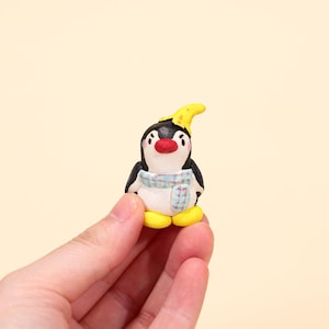Könnte beinhalten: Kleine, handgefertigte Pinguinfigur. Der Pinguin hat einen schwarzen Körper, einen weißen Bauch und einen roten Schnabel. Er trägt einen blau-weißen Schal und einen gelben Hut. Der Pinguin steht auf gelben Füßen.