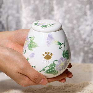 May include: White ceramic pet urn with a lid, adorned with hand-painted purple morning glories and green leaves. A gold paw print and the name "Pip" are featured on the urn, which is held in someone's hands.
