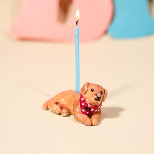 May include: A brown dog-shaped birthday candle holder with a red bandana featuring white hearts. A lit blue candle is inserted into the dog's back. The background is a light beige with blurred pink and blue shapes.