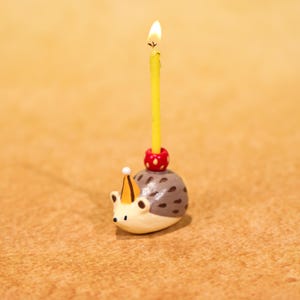Puede incluir: Un caprichoso portavelas de cerámica con forma de erizo con una vela amarilla encendida. El erizo está pintado con detalles marrones, blancos y grises, con un gorro de fiesta. La vela se sujeta con un anillo rojo y blanco con lunares.