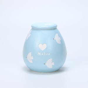 Puede incluir: Urna de cerámica azul claro con tapa, decorada con palomas blancas y un corazón blanco. El nombre "Malou" está escrito en cursiva debajo del corazón. La urna tiene una forma redondeada, en forma de huevo.