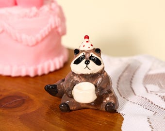May include: A small ceramic raccoon figurine featuring brown and white fur, black eye patches, and a pointed party hat with red polka dots. The raccoon is sitting in a seated position, holding a small white cup with both front paws.