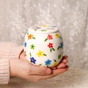 May include: A white ceramic jar with a lid, adorned with colorful hand-painted flowers in red, yellow, blue, and orange. The jar is held in open hands, showcasing its round shape and floral design. The jar is approximately 4 inches in diameter.