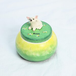 May include: A ceramic jar with a green and yellow gradient design. The lid is green with a white dog figurine and the word "Prinz". The lid is decorated with small white and yellow flowers.