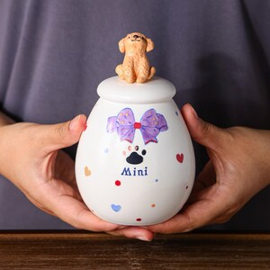 Pode incluir: Um pote de cerâmica branco com tampa, decorado com um laço roxo, uma pegada e corações coloridos. Uma pequena figura de cachorro marrom claro está no topo da tampa. A palavra "Mini" está impressa no pote.
