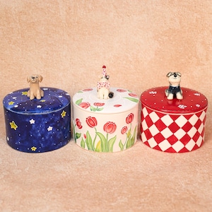 May include: Three decorative ceramic jars with lids. One jar is navy blue with a starry night design and a dog figurine. Another is white with red tulip flowers and a cat figurine. The third is red and white checkered with a dog figurine.