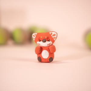 May include: A handcrafted, orange-red fox figurine with a white belly and accents. The fox has closed eyes, a black nose, and brown feet. The ears have white and orange patterns. The figurine is set against a soft pink background.
