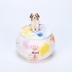 May include: A white ceramic jar with a lid featuring colorful paint splatters and a small dog figurine on top. The jar has the word "Mini" and a paw print design on the front.