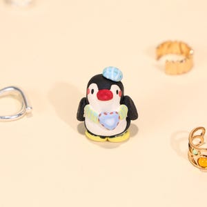 Könnte beinhalten: Handgefertigte Pinguin-Figur mit schwarzem Körper, weißem Bauch und gelben Füßen. Der Pinguin trägt einen blauen Hut und Schal und hält ein Herz. Der Hintergrund ist hellbeige, mit Ringen.