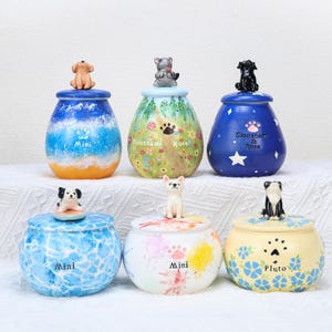 May include: Six decorative ceramic jars with lids, each topped with a dog figurine. The jars feature various designs, including ocean waves, floral patterns, and star motifs. Some jars have the word "Mini" or a dog's name printed on them.