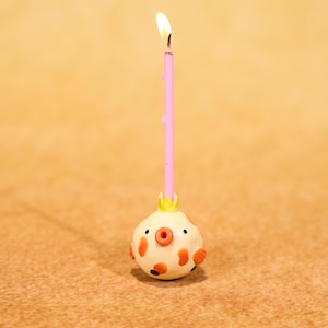 May include: A lit pink birthday candle with a yellow flame sits atop a small, round, cream-colored figure with orange and black details. The figure has a small yellow crown. The candle is on a brown surface.