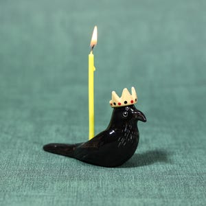 Pode incluir: Um castiçal preto em forma de corvo com uma pequena coroa, segurando uma vela amarela acesa. O corvo tem um acabamento brilhante e está sobre um fundo verde-azulado. A coroa é branco-sujo com detalhes vermelhos.