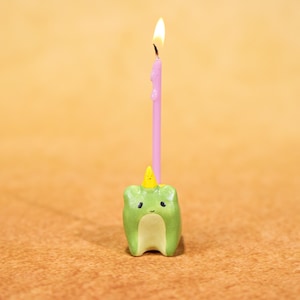 May include: A novelty birthday candle. The base is a green tooth shape with a yellow horn and a smiling face. A pink candle is lit, with wax dripping down. The background is a warm, solid colour.
