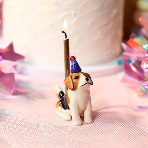Puede incluir: Un portavelas de cumpleaños de perro beagle de cerámica con un gorro de fiesta azul y una vela dorada encendida. El perro es blanco, marrón y negro, sentado frente a una tarta glaseada blanca. El fondo incluye decoraciones rosas y moradas.