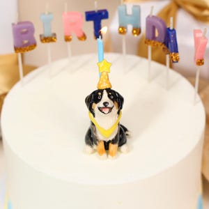 May include: A birthday cake with a decorative dog figurine and candles spelling "BIRTHDAY!". The dog figurine is black, white, and brown, wearing a yellow bandana and party hat. A lit blue candle with a star topper is in the center.