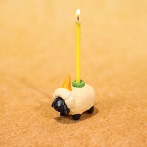 Puede incluir: Una vela de cumpleaños caprichosa con una figura de oveja y una vela amarilla. La oveja tiene un cuerpo color crema, cara y patas negras, y un pequeño cuerno naranja. La vela está encendida con una pequeña llama.