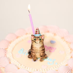 May include: A decorated birthday cake featuring a cat candle. The candle is a tabby cat wearing a party hat, with a lit pink candle. The cake has pink frosting and the word "Birthday" written on it.
