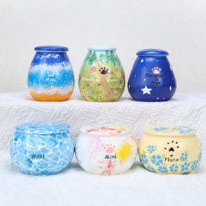 May include: Six decorative ceramic pet urns in various colors and designs. The urns feature paw prints, floral patterns, and text such as "Mini" and pet names. The urns are in shades of blue, yellow, and white.