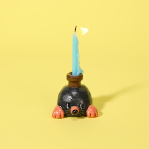 May include: A whimsical candle holder shaped like a mole, with a black body, pink paws, and a brown neck. A blue candle is lit, with a yellow flame. The candle holder is on a yellow background.
