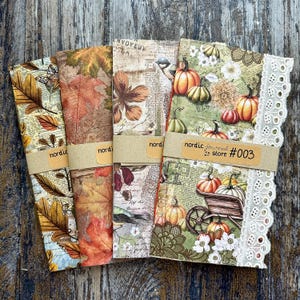 Könnte beinhalten: Vier dekorative Journale mit herbstlichen Einbänden. Die Einbände zeigen Bilder von Blättern, Kürbissen und floralen Mustern. Jedes Journal ist mit einem braunen Papierband gebunden, auf dem "nordic journal store #003" steht. Das rechte Journal hat eine weiße Spitzenborte.