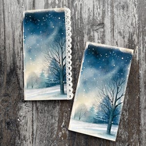 Könnte beinhalten: Zwei Aquarellmalereien zeigen eine verschneite Winterszene mit kahlen Bäumen und einem Wald. Die Gemälde zeigen einen dunkelblauen Himmel mit fallendem Schnee und eine weiße Spitzentrimmung auf einem. Das Kunstwerk befindet sich auf einer Holzoberfläche.