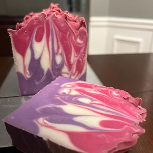 May include: A bar of handmade soap with swirls of pink, white, and purple. The soap is cut into two pieces, one of which is partially visible.