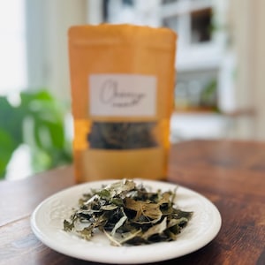Peut inclure: Une petite assiette blanche à bord décoratif contient une pile de feuilles vertes séchées. En arrière-plan, un sachet en papier brun avec une étiquette blanche et le mot "Chancy" suggère un produit à base de thé ou de plantes.