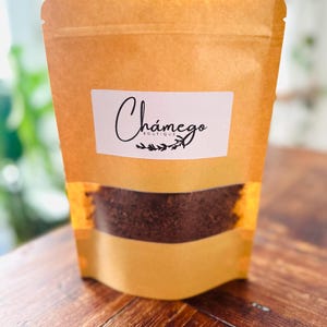 Puede incluir: Una bolsa de papel marrón con una etiqueta que dice "Chámego Boutique" y contiene hojas de té marrón sueltas.