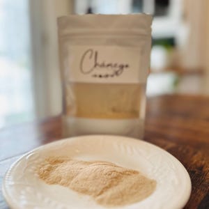 May include: A white ceramic plate displays a pile of light brown powder. Behind it, a clear bag with the word "Chamego" in cursive script is visible. The bag is filled with a similar powder.