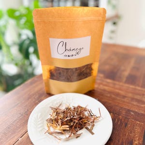 以下が含まれることがあります： 白いラベルに「Chámego」と小さな葉の模様が描かれた茶色の紙袋。袋の中には、茶色の乾燥ハーブが緩く入っています。小さな白い皿に、茶色の乾燥ハーブが山盛りになっています。