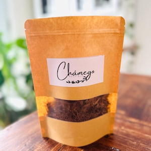 以下が含まれることがあります： 白いラベルに「Chámego Boutique」と書かれた茶色の紙袋に、乾燥ハーブが入っています。