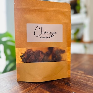 May include: A brown paper bag with a clear window showing dried tea leaves. The bag features a white label with the cursive text "Chámego" and a small botanical design. The bag is on a wooden surface.