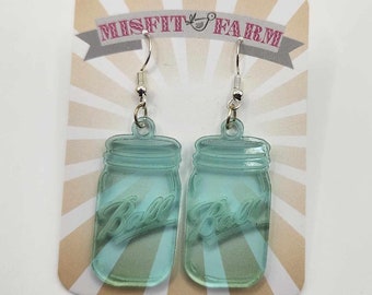 May include: A pair of light blue glass jar earrings with silver hooks. The jars have the word "Ball" embossed on them. The earrings are on a white card with the words "Misfit Farm" printed on it.