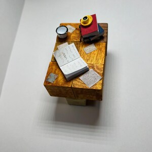 May include: A miniature wooden table with a stack of books, a small cup, and a notebook with handwritten notes. The table is made of multiple wooden blocks and has a rustic, handcrafted look.